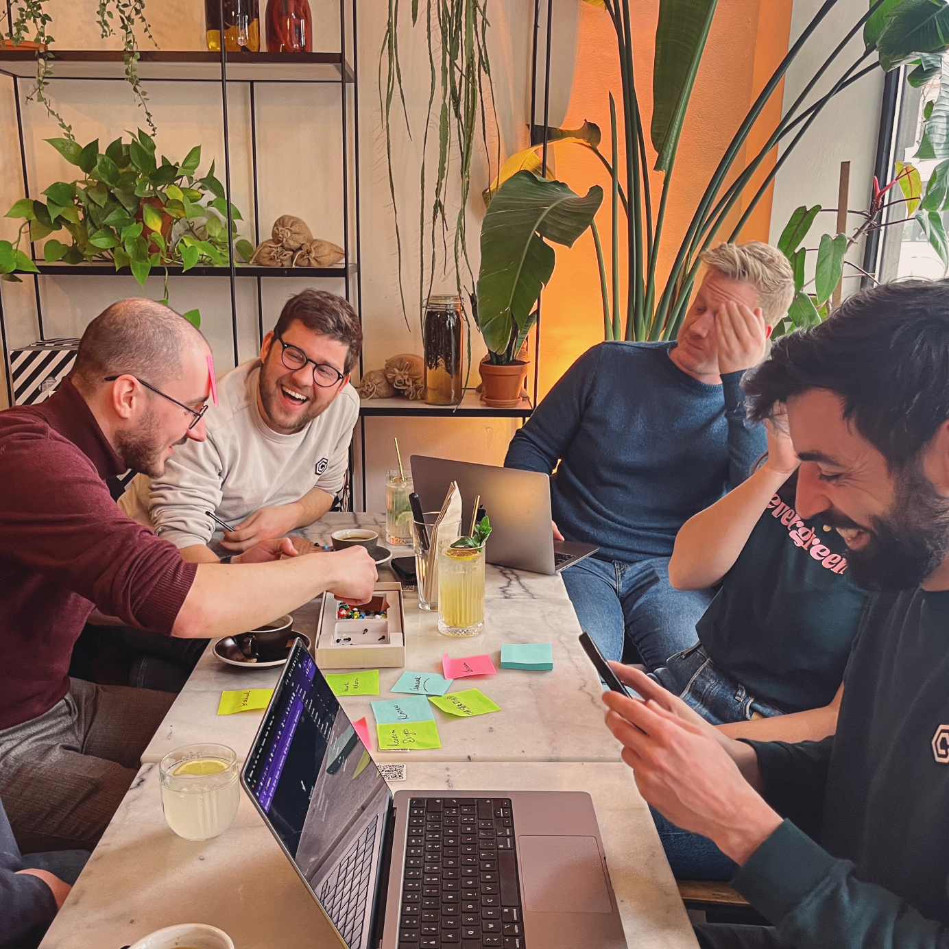 Five colleagues sitting together at a table, one is looking at his screen, the other four are playing and laughing together